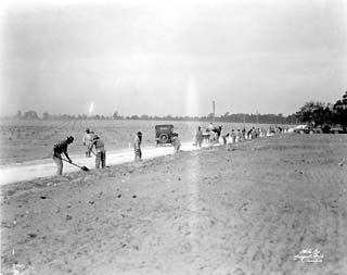 Open stretch of road lined with men in prison uniforms using shovels.