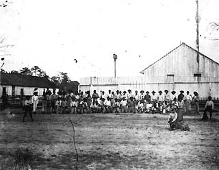 Convicts lined up in front of large wooden buildings.