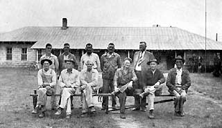 Eleven men sitting on wooden bench in front of block building with tin roof.