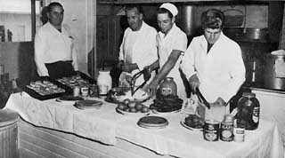 Four men, three in chief's uniforms, preparing a table of food.