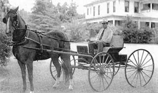 Man sitting in horse drawn buggy in front of two story wood house.