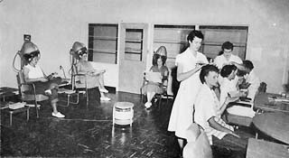 Three women sitting under hair dryers, two beautitions cutting har on two other women.