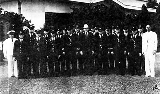 Approx. 20 men in dark uniforms, two in white, and person in center has white hat.