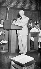 Man in light suit standing inside chapel at podium.