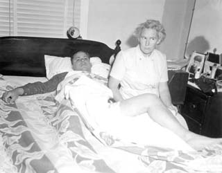 Wainwright laying on back in hospital bed with woman sitting on left.