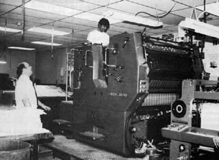 Officer standing next to large printing machine while inmate works near top.