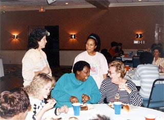 Seven women sitting at/standing near table with food.